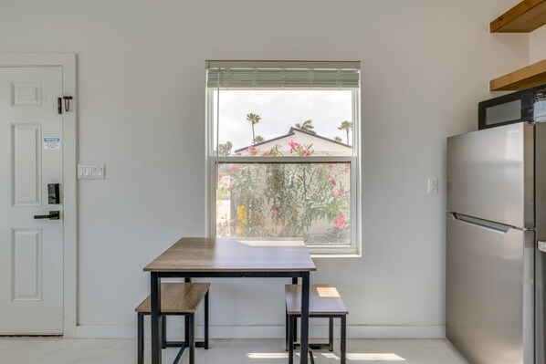 Une cuisine équipée d’un réfrigérateur encastré, une fenêtre donnant sur une maison et des palmiers, et une table avec deux chaises.
