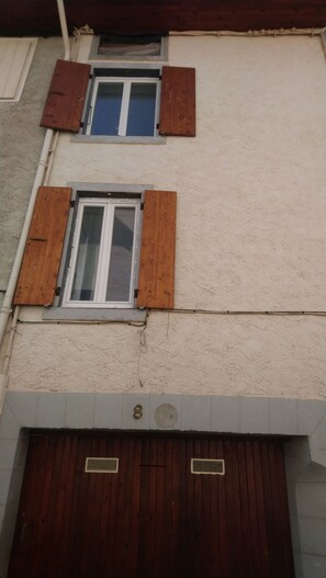 Un bâtiment doté de deux fenêtres, chacune avec des volets en bois, et d’une porte de garage sur laquelle est apposée une plaque d’immatriculation.