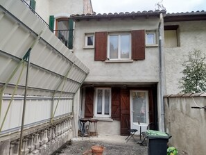 Ein zweistöckiges Haus mit einem kleinen Balkon, einer Tür mit Fenster und einem Tisch mit Stuhl davor.