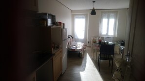 Et kompakt køkken med et køleskab, en mikrobølgeovn og en vask. Der er et spiseområde med et bord og stole, og et vindue giver naturligt lys.