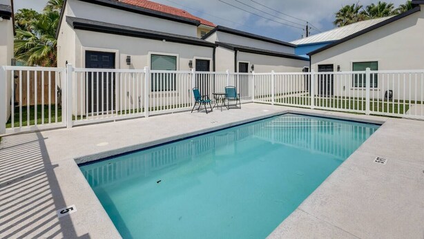 A swimming pool with a white fence, two chairs, and a building with a red roof. - Poolside Studio on Padre (#2) (South Padre Island)