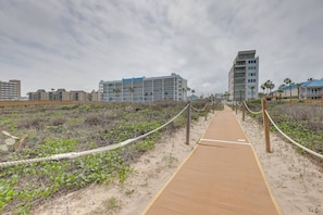 A sandy pathway leading to a beachfront hotel with multiple floors and balconies.