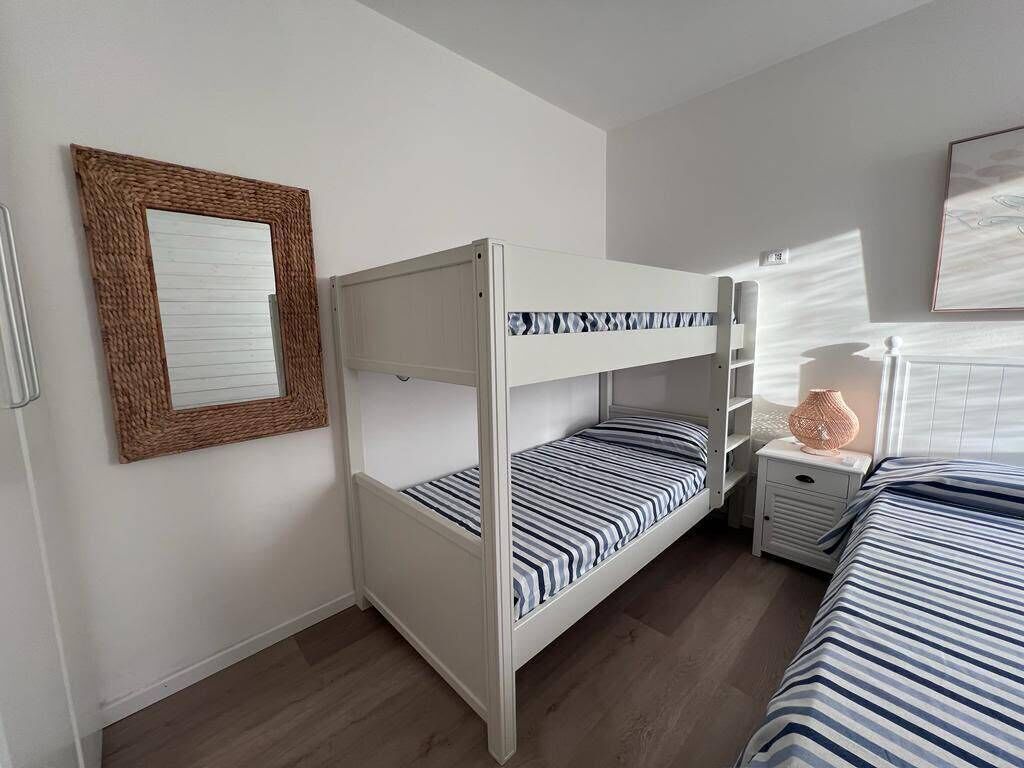 Habitación con literas de estructura blanca, una mesita de noche con lámpara y un cuadro en la pared.