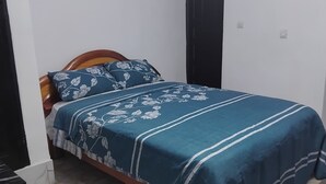 Um quarto com uma cama, uma mesa de cabeceira, uma prateleira na parede com objetos decorativos e uma porta.