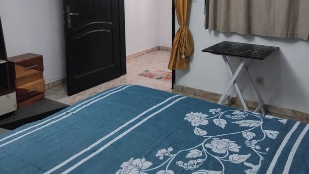 Um quarto com uma cama, uma porta, uma cortina, uma mesinha e um lustre.