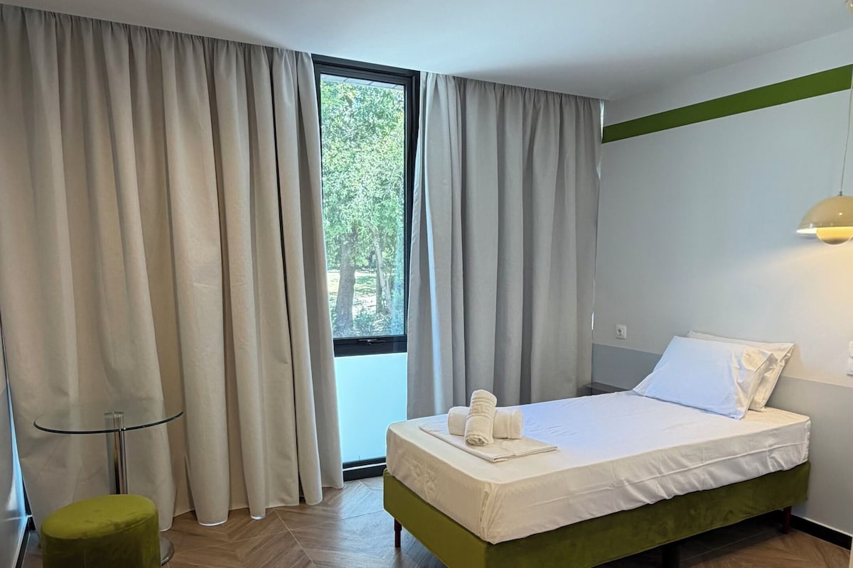 Quarto de hotel com cama, cortinas, janela, um banquinho verde e piso com padrão losango.