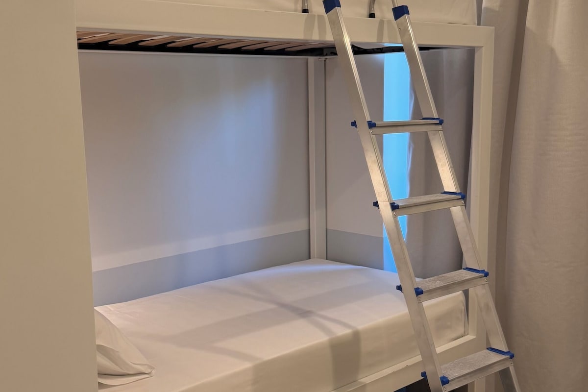 Uma cama beliche com escada, uma faixa verde na parede e uma janela com cortinas.