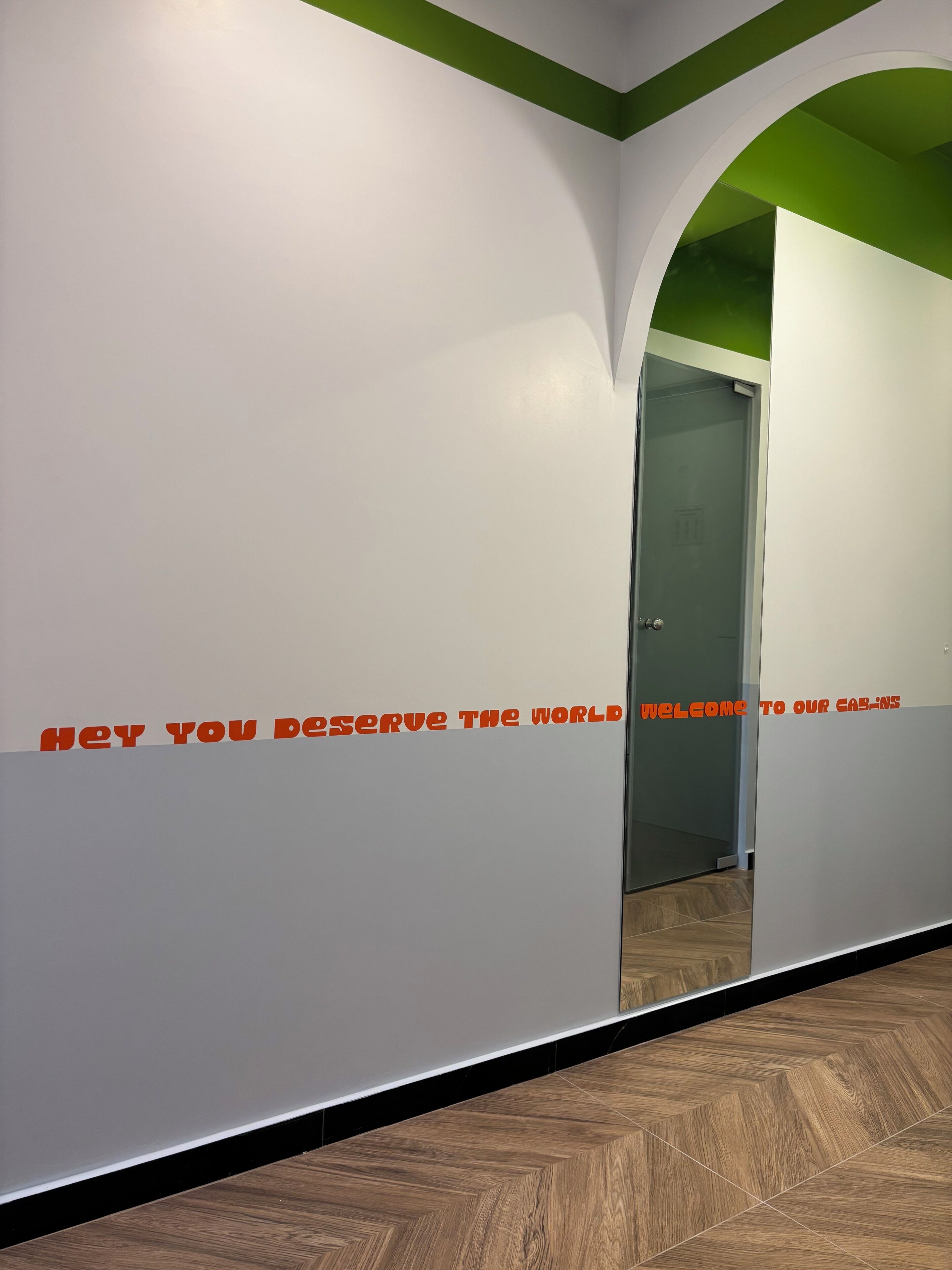 Ein Flur mit einer Spiegelwand und einem grünen Bogen. Auf der Wand steht der Text 'HEY YOU DESERVE THE WORLD WELCOME TO OUR CABINS'.