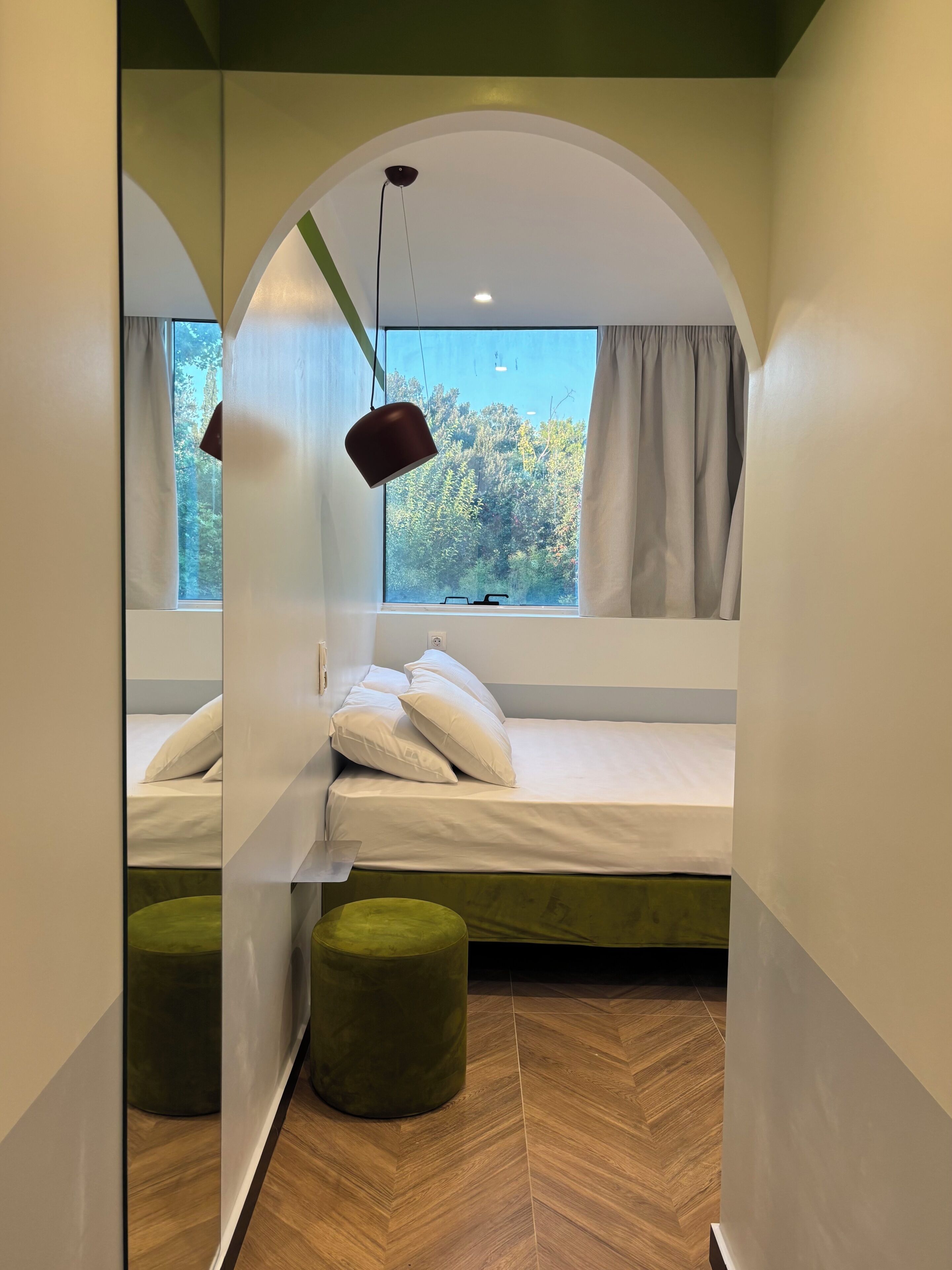 Un dormitorio con cama, lámpara colgante, ventana con cortinas y vista a árboles.