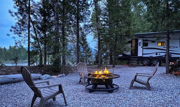 Un camping con un pozo de fuego, dos sillas Adirondack y una caravana.