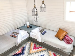 A cozy seating area with a bench and two small beds, a patterned rug, and three hanging light fixtures.