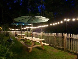 Un área de patio al aire libre con mesas de picnic y un gran paraguas, rodeada por una valla de madera y luces colgantes.