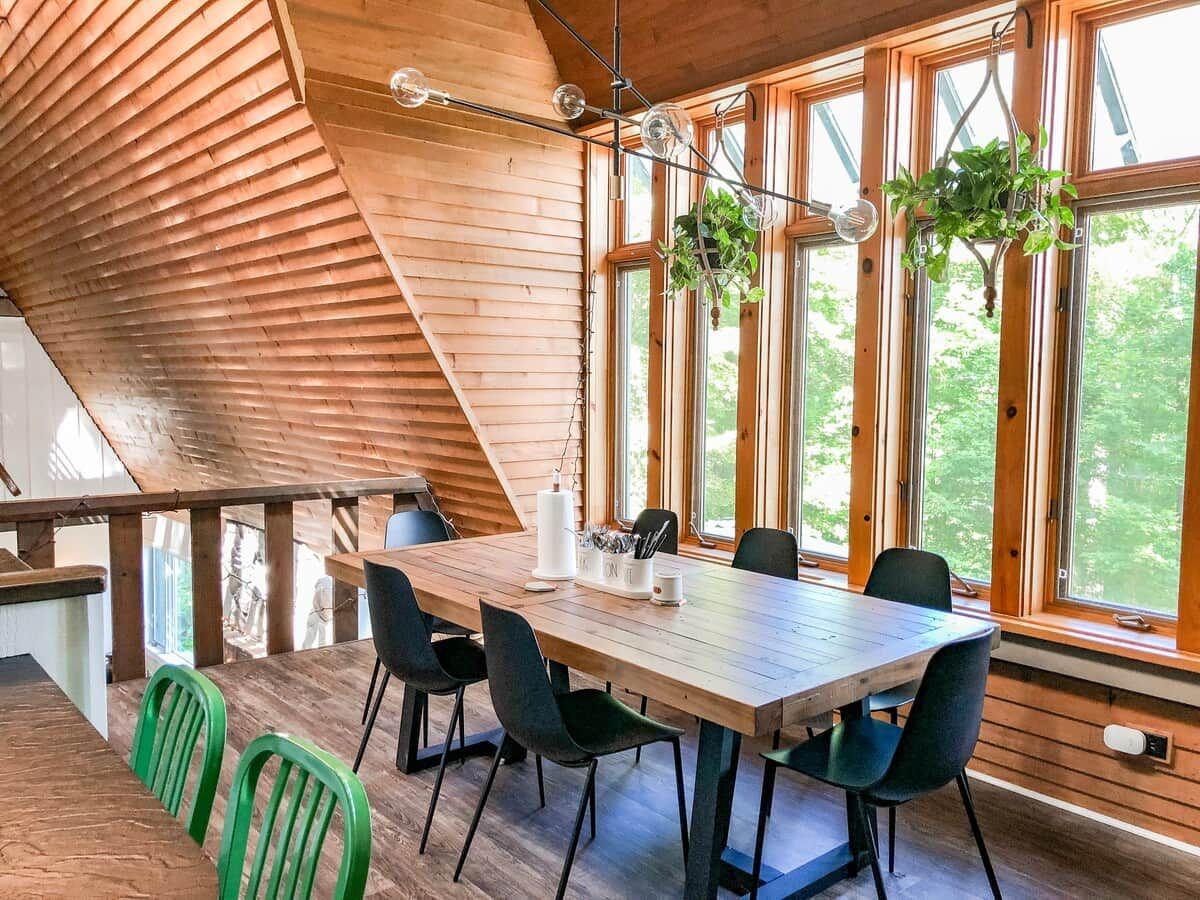 Un coin repas en bois, avec une table et des chaises, de grandes fenêtres et des plantes suspendues.