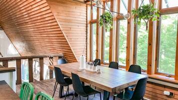 Un coin repas en bois, avec une table et des chaises, de grandes fenêtres et des plantes suspendues.