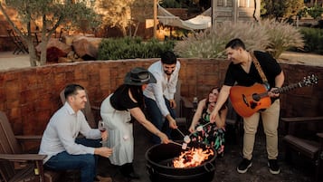 Um grupo de pessoas reunido em volta de uma fogueira, com um músico tocando violão, sentadas em bancos de madeira, em ambiente externo com paredes de pedra e plantas.