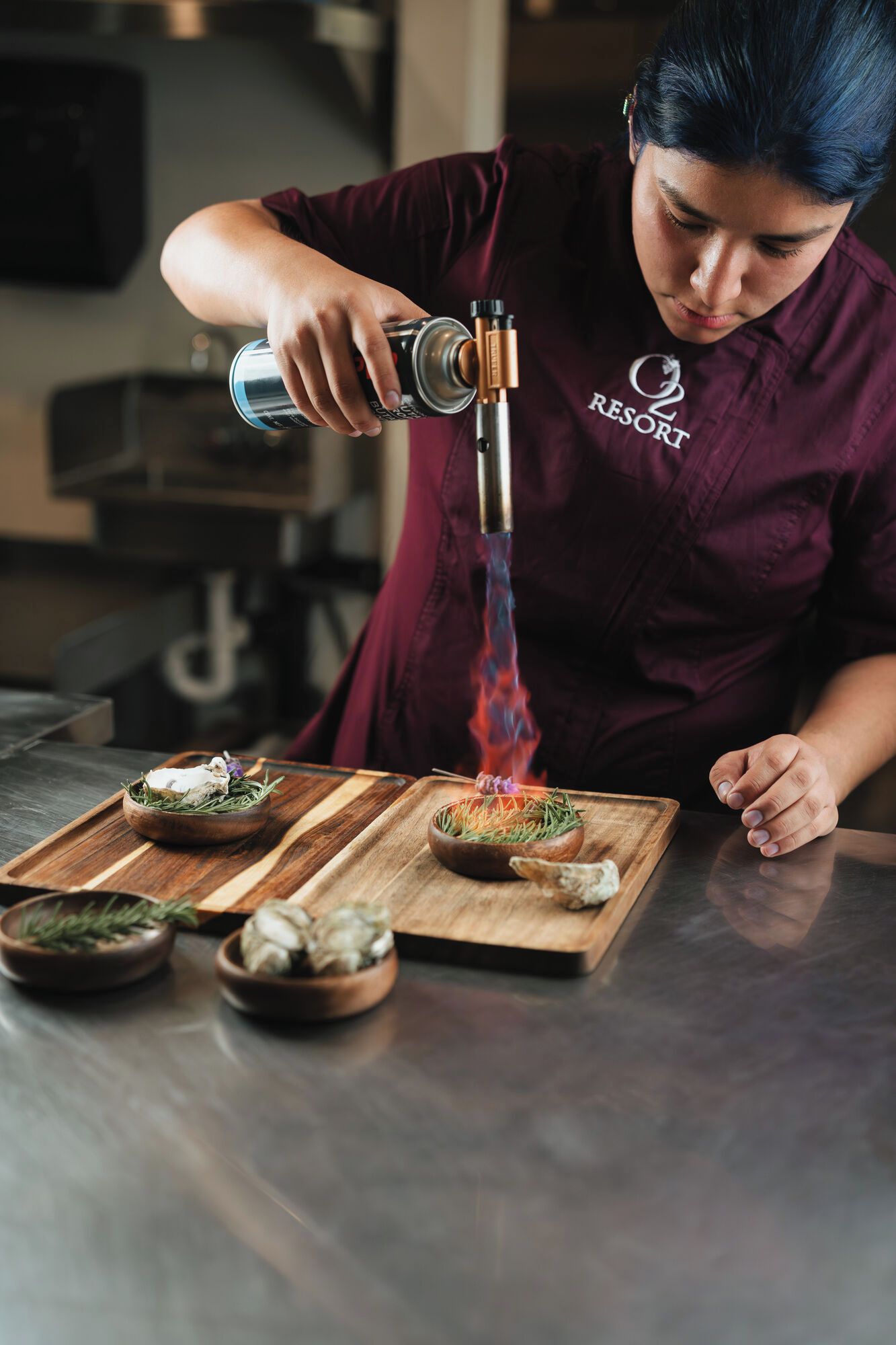 A person in a maroon shirt with a logo, pouring a liquid from a can into a device that emits blue flames.
