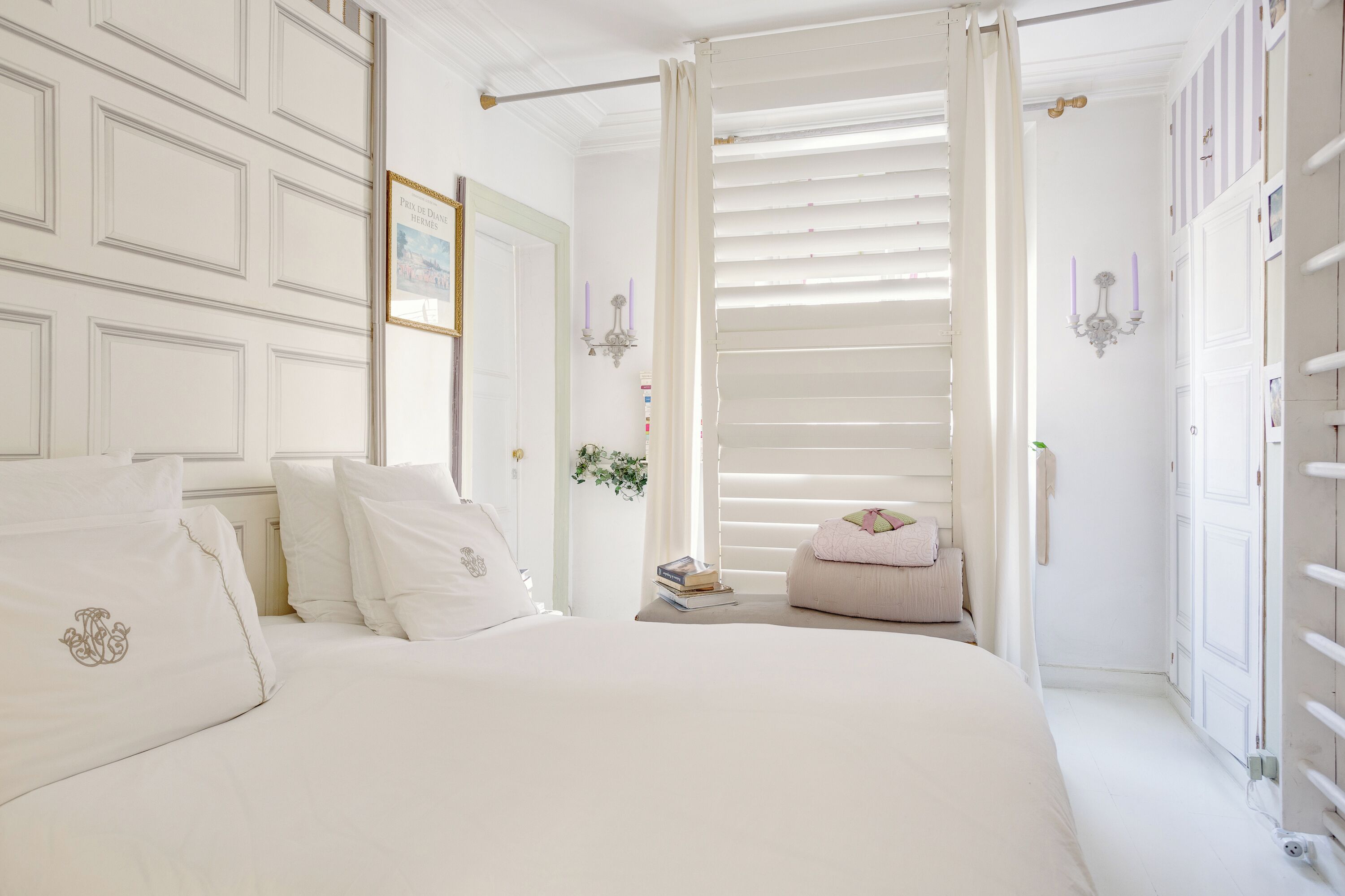 A bedroom with a large bed, a bedside table with books, a hanging plant, and a wardrobe with a mirror.