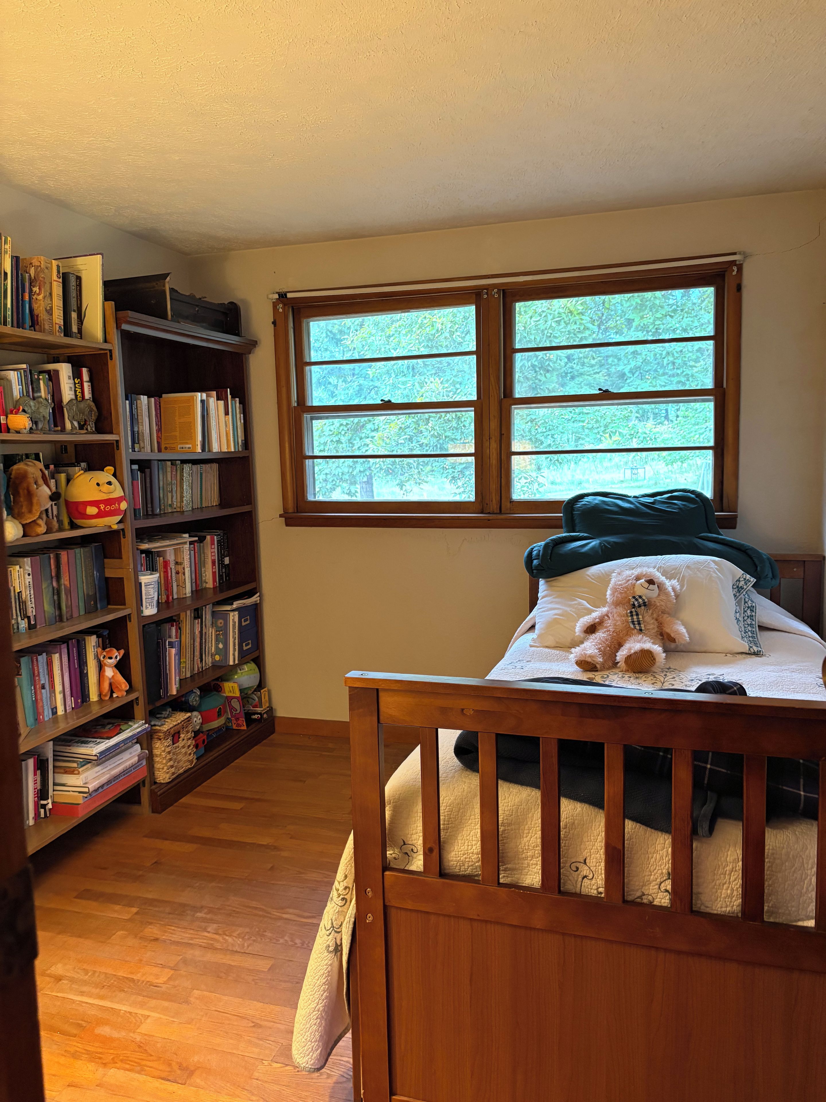 Una habitación con una cuna de madera, una estantería llena de libros y juguetes, y un ventanal con vista a zonas verdes.