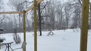 Una estructura de pérgola de madera con una silla metálica cubierta de nieve, una mesa y un patio nevado con árboles al fondo.