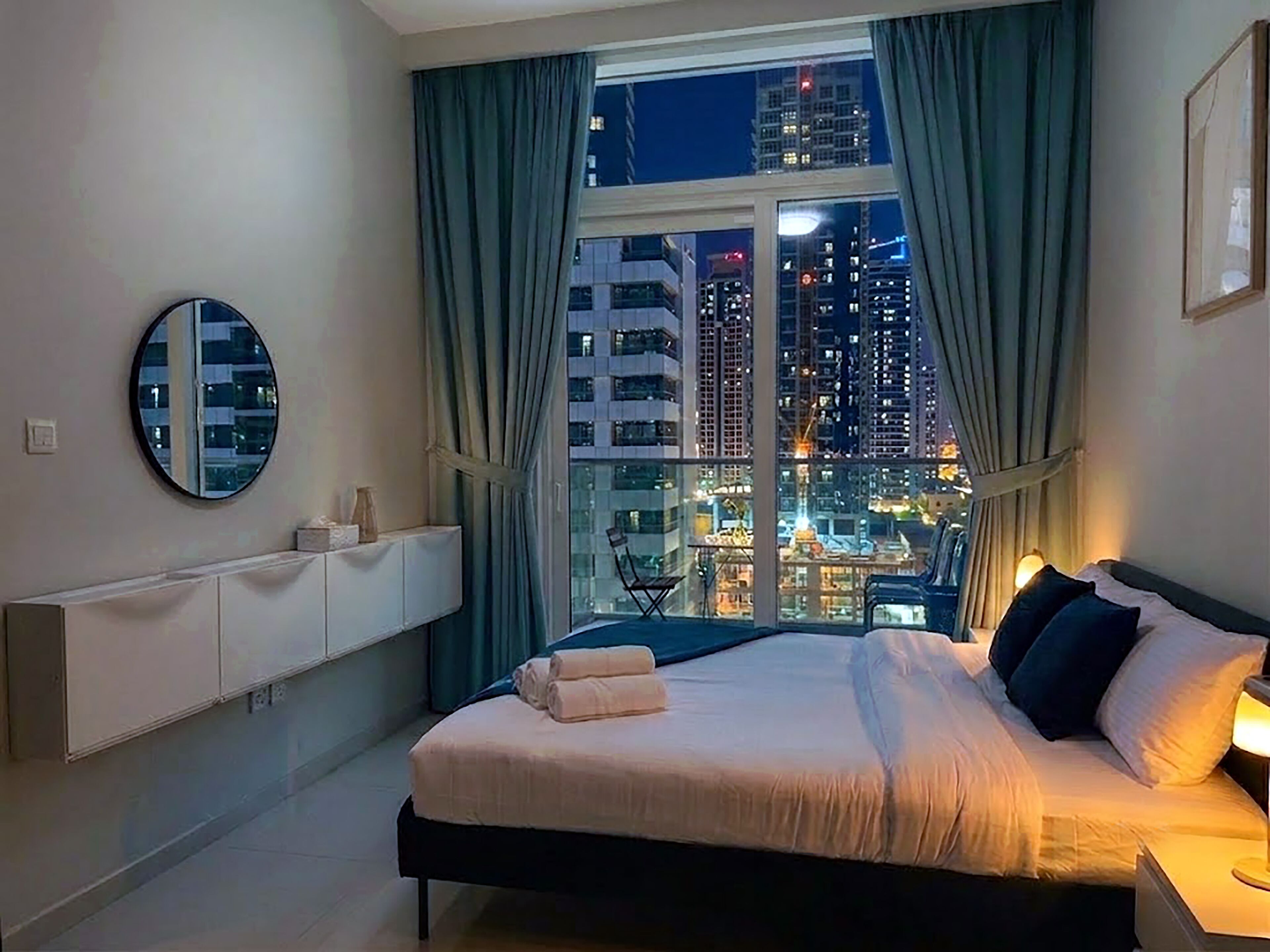 Une chambre d’hôtel avec un lit, une table de chevet, un miroir et une vue sur le paysage urbain illuminé la nuit.