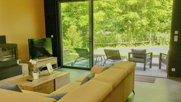 Uma sala de estar moderna com um sofá, uma televisão e vista para o verde através de grandes janelas.