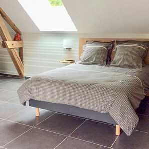 A modern bedroom with a sloped ceiling, a bed with patterned bedding, a bedside table with a lamp, and a view of greenery outside. - Charming cottage with sauna near Cambrai (Walincourt-Selvigny)