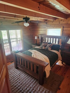 Ein Schlafzimmer einer Holzhütte mit Bett, Nachttisch, Lampe und einem Fenster mit Jalousien.