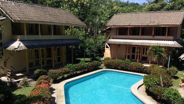 A swimming pool surrounded by lush greenery and two wooden buildings with tiled roofs. - Villas de la Bahía Playa Tambor (Cobano)
