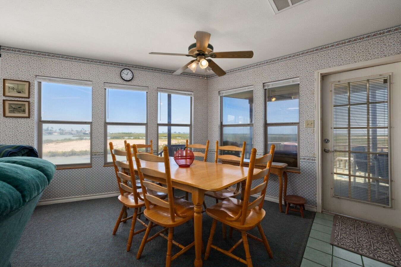 A dining room with a wooden table and chairs, a ceiling fan, and large windows with a view of the beach.