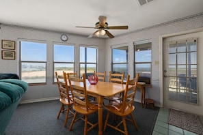 A dining room with a wooden table and chairs, a ceiling fan, and large windows with a view of the beach.