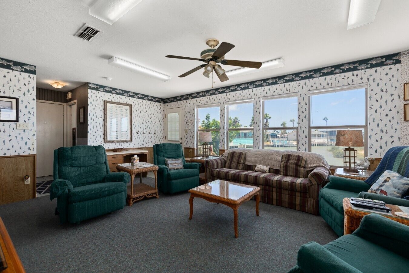 A living room with a ceiling fan, large windows, and a view of the outdoors.