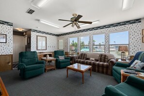 A living room with a ceiling fan, large windows, and a view of the outdoors. - Oyster Cove in Long Island Village (Port Isabel)