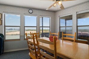 A room with a wooden dining table, four chairs, a ceiling fan, and large windows offering a view of the beach. - Oyster Cove in Long Island Village (Port Isabel)