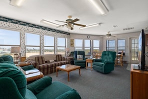 A spacious living room with a large sofa, two armchairs, a coffee table, and a television. There are multiple windows providing a view of the outdoors.
