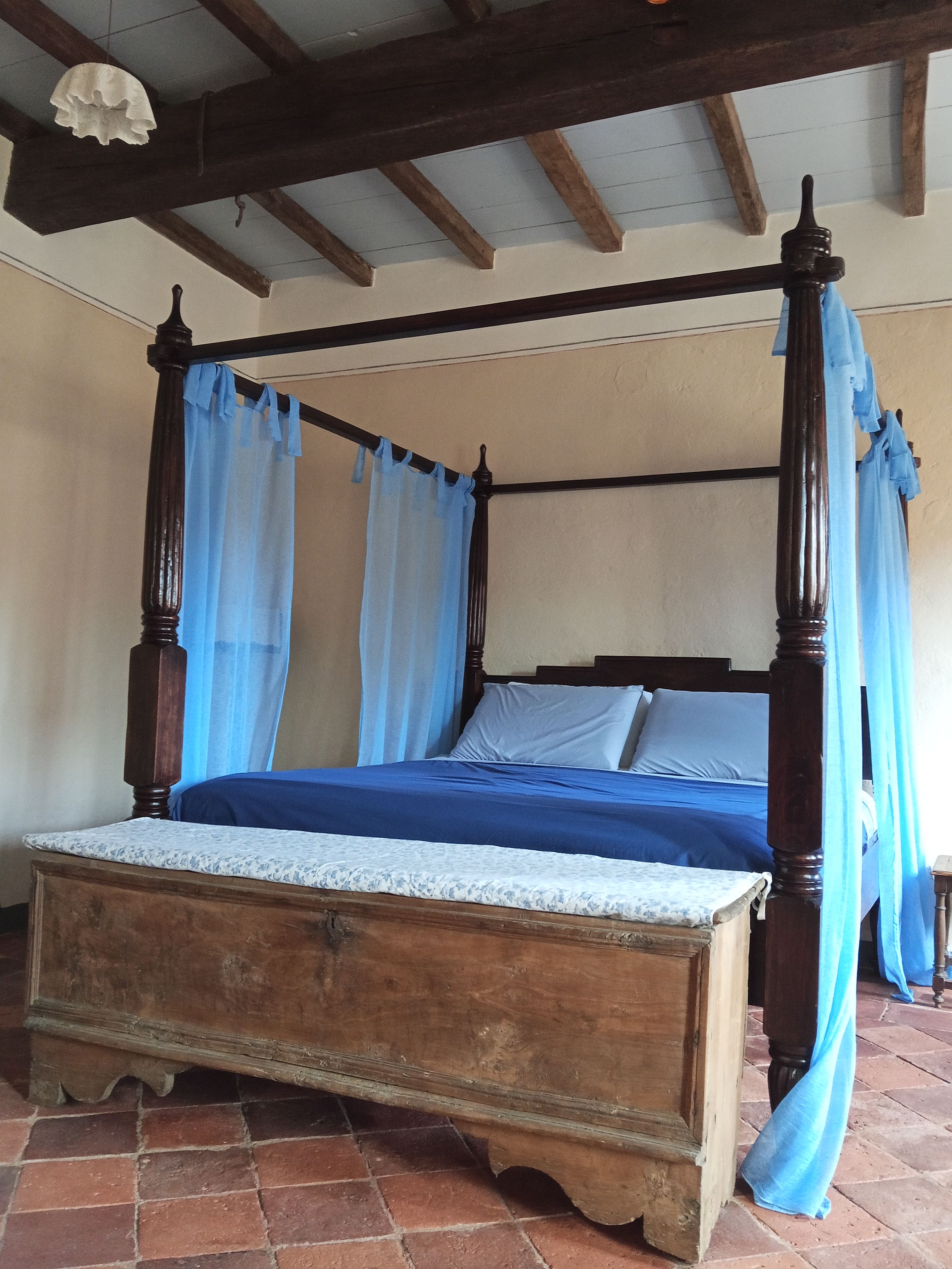Un letto a baldacchino con struttura in legno e tendaggi blu, posto su un pavimento piastrellato sotto un soffitto spiovente.