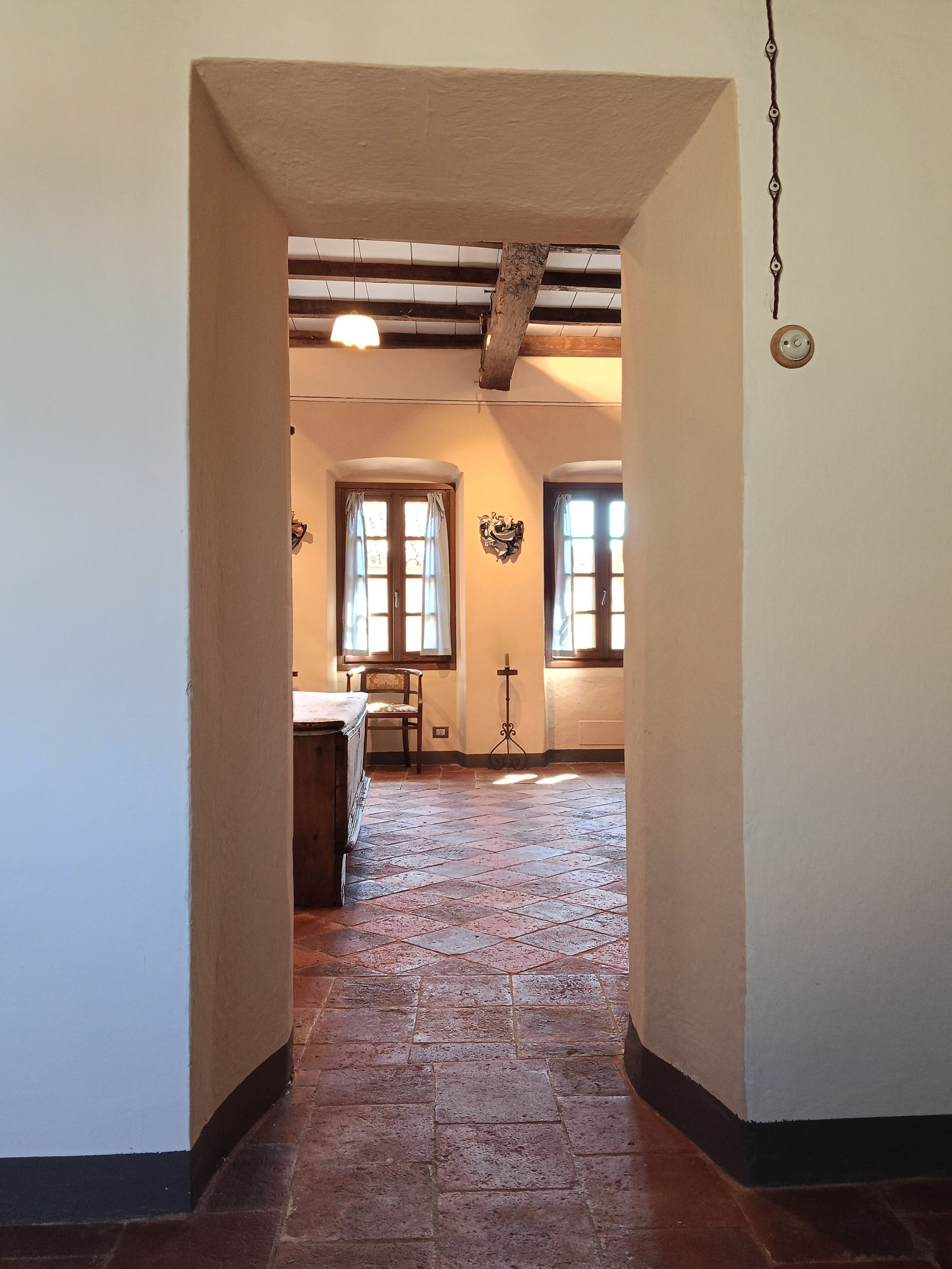 A hallway with a tiled floor, wooden beams on the ceiling, and two windows with curtains.