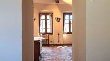 A hallway with a tiled floor, wooden beams on the ceiling, and two windows with curtains.