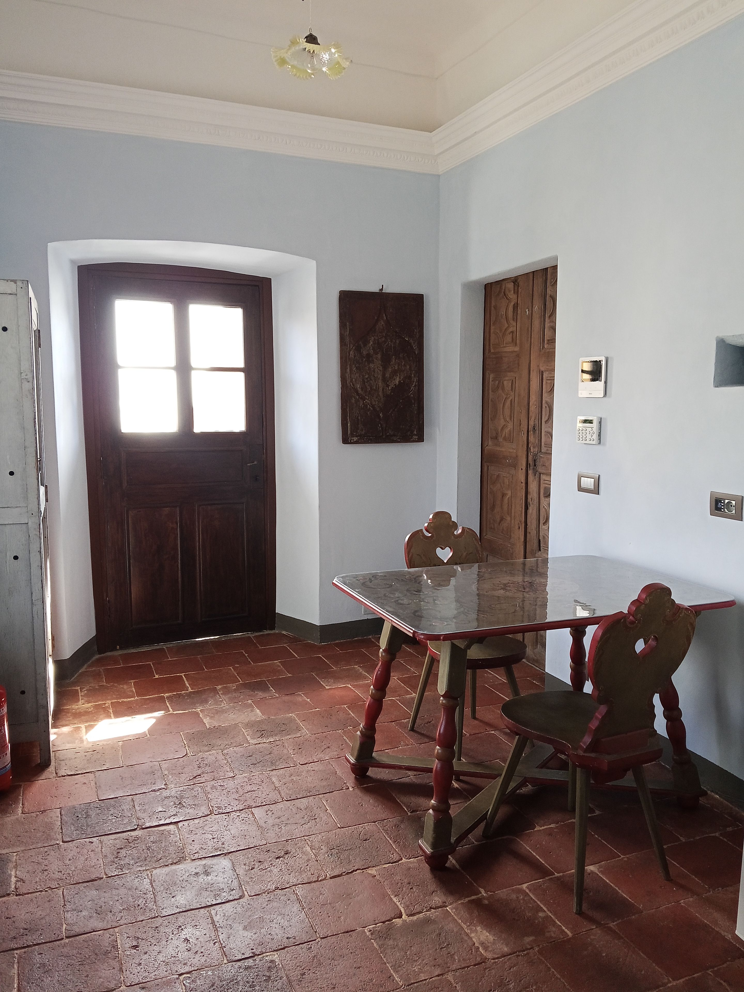 Een kamer met een houten deur, een glazen tafel en een stoel. Er zijn twee lichtschakelaars aan de muur en een klein plankje.