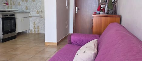 Una habitación con un sofá púrpura, una zona de cocina con armarios y una estufa, y una puerta de madera.