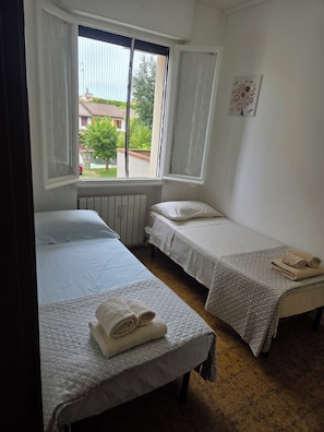 Habitación con dos camas individuales, un radiador, ventana con cortinas y vistas a edificios y árboles.