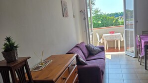 A room with a wooden dresser, purple sofa, and a dining table with chairs. There is a balcony with a view of greenery. - Apartment 300 meters from the beach, with air conditioning and a private parking space (Lido Adriano)