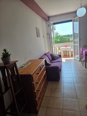 Ein Zimmer mit einem Holzkoffer, einem violetten Sofa und einem Esstisch mit Stühlen. Ein Balkon bietet Blick auf Grünflächen.