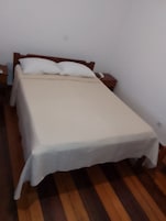 Uma cama de solteiro com lençol branco e duas almofadas, um criado-mudo de madeira com um telefone, e piso de madeira.