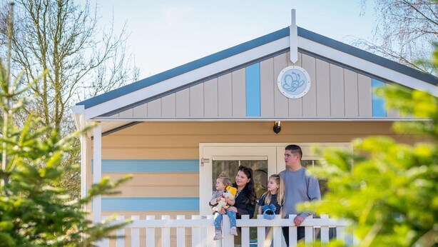 A family standing at a house's porch with a white railing, a blue door, and a circular emblem on the wall. - Family Holiday Home in Resort in Hoogerheide (Hoogerheide)