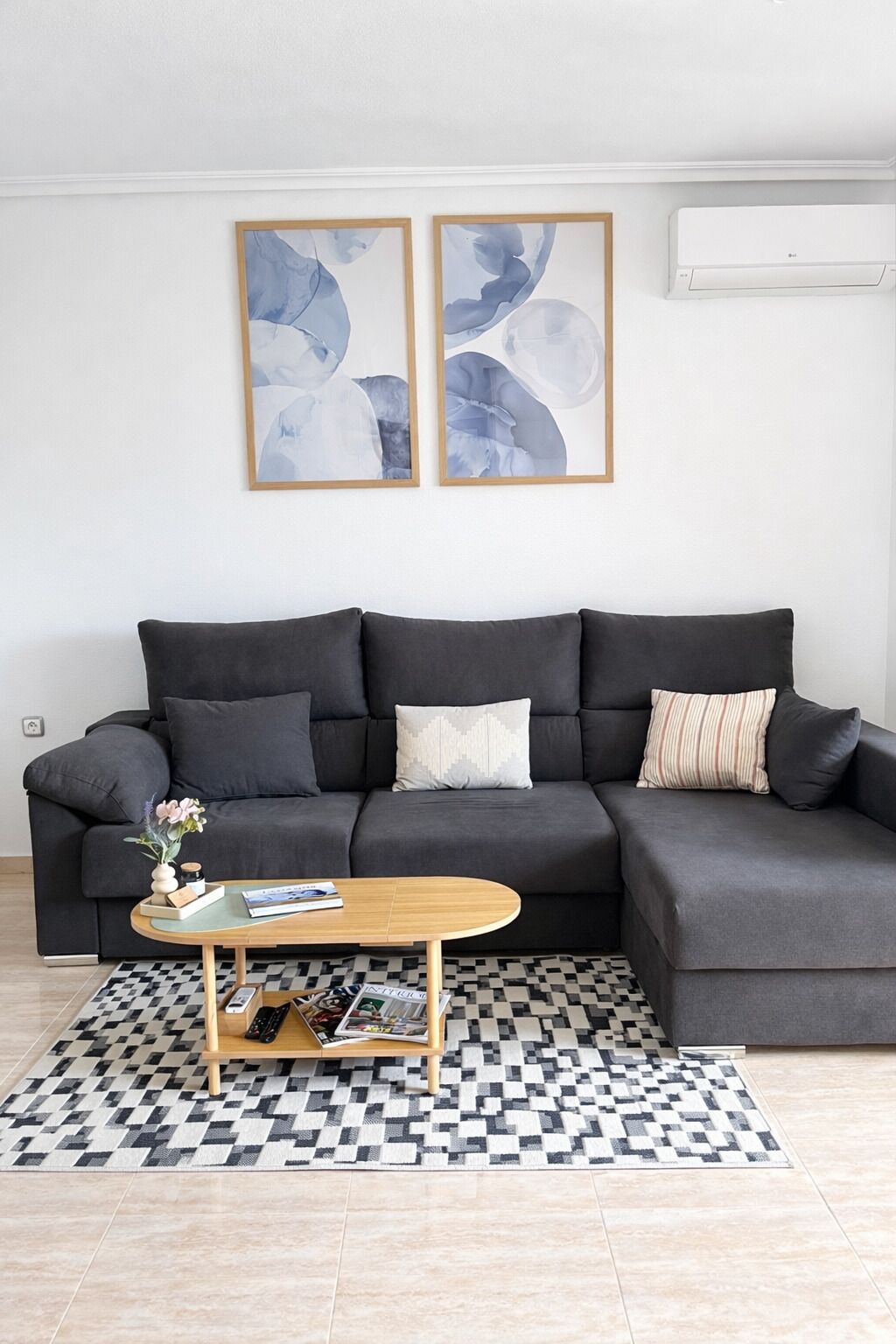 Un salon moderne avec un canapé d’angle gris, une table basse en bois et un tapis à carreaux. Deux tableaux abstraits sont accrochés au mur au-dessus du canapé.