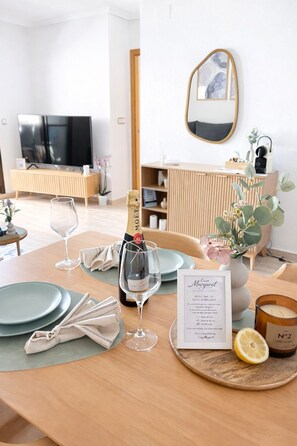 Mesa de jantar posta com pratos, copos, uma garrafa de champanhe, uma vela e um cardápio em um quarto moderno, bem iluminado, com televisão e espelho na parede.