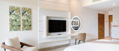 A modern hotel room with a flat-screen TV mounted on a white wall, a round mirror, a wooden chair, and a bed with a white headboard.