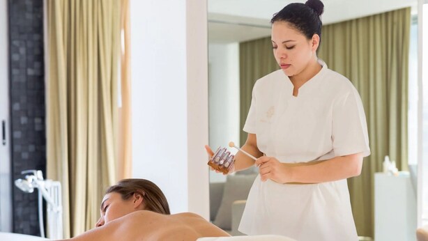 En person ligger på en seng i et rum med en massagestol, en massør står ved siden af, og et massagestue står i baggrunden.