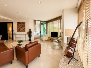 A modern living room with a brown sofa, a glass coffee table, and a guitar leaning against a wall. - Fully Seaview 2BR Apartment, Walk to Kamala Beach (Kamala)