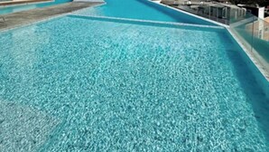 Ein Schwimmbecken mit klarem, blauem Wasser, umgeben von einem weißen Gebäude mit Balkonen, vor einer Berglandschaft und einem wolkenlosen Himmel.